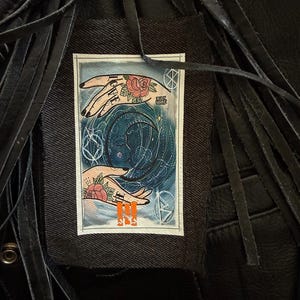 Può includere: Una toppa in denim nero con un disegno di tarocchi raffigurante due mani, rose e la scritta "LOVE". La toppa è cucita su una giacca di pelle nera con frange.