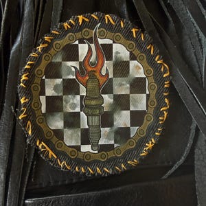 Può includere: Toppa rotonda con un design di candela infuocata su sfondo a scacchi bianco e nero. La toppa ha un bordo a catena e cuciture gialle. Su pelle nera con frange.