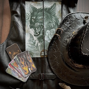 Può includere: Gilet in pelle nera con grafica di lupo sul retro, cappello da cowboy nero e mazzo di tarocchi con disegni di fiamme. Il gilet ha una chiusura con fibbia. Il cappello ha un design cucito e due anelli dorati.