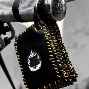 May include: A black leather keychain tag featuring a white flame graphic on the front. The rectangular tag is constructed from layered leather pieces joined by thick, visible yellow stitching along the edges and hangs from a metal ring.