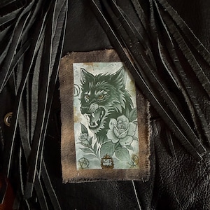 Può includere: Giacca di pelle nera con lunghe frange. Una toppa rettangolare è cucita sulla giacca, con un'illustrazione dettagliata di una testa di lupo, rose e la scritta "RUST NOIRE". L'opera è nei toni del verde e del grigio.