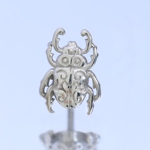 May include: A silver-toned stud earring featuring a detailed beetle design with intricate embossed scrollwork patterns on its back. The beetle has prominent mandibles and six legs, attached to a straight post with a matching silver-toned backing.