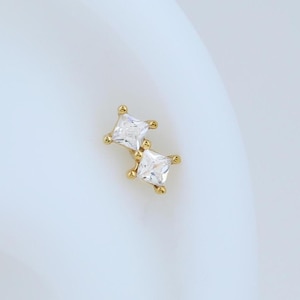 May include: A small, gold-colored earring with two square, clear gemstones. The earring is set against a white background, highlighting the jewelry's design and sparkle. The earring is a stud style.