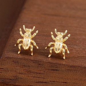 May include: A pair of gold-colored stud earrings shaped like small beetles. The earrings are detailed with legs, antennae, and body segments. They are displayed on a dark wooden surface.