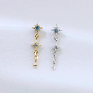 May include: Two dangling earrings featuring a starburst design with small blue stones. One earring is gold-toned and the other is silver-toned. Each piece consists of two star-shaped charms connected by a delicate chain, ending in a tiny star point.