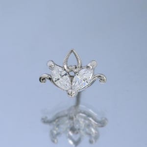 Könnte beinhalten: Ein silberner Ohrstecker mit floralem Design. Der Ohrring hat drei klare, marquiseförmige Steine in der Mitte, umgeben von filigranen, gebogenen Metallarbeiten. Der Ohrring wird auf einer reflektierenden Oberfläche präsentiert.