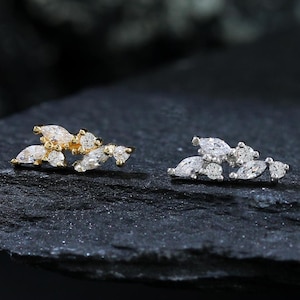 May include: A pair of gold-toned and silver-toned stud earrings. Each earring features a cluster of small, marquise-cut gemstones. The earrings are displayed on a dark, textured surface, with a blurred dark background.