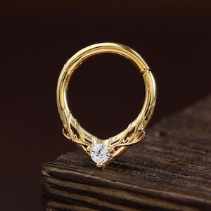 Puede incluir: Un anillo de tabique dorado con un delicado diseño en forma de cuerno. El anillo presenta una pequeña gema transparente en el centro, añadiendo un toque de brillo. La joya se exhibe sobre una superficie de madera oscura.