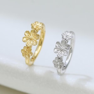 Op de afbeelding: Twee ringen met bloemmotieven. Eén ring is goudkleurig, met drie kleine bloemen. De andere ring is zilverkleurig, eveneens met drie bloemen. Beide ringen worden gepresenteerd tegen een witte achtergrond.