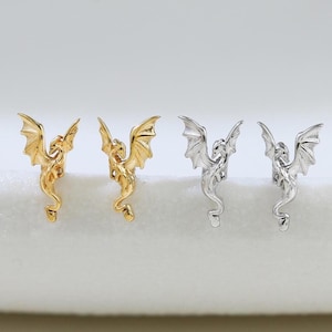 May include: A set of four stud earrings shaped like small dragons. Two earrings are gold-toned and two are silver-toned. Each dragon is depicted with wings spread upwards, a long tail, and detailed features, standing upright in a vertical orientation.