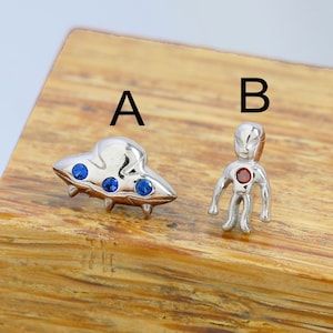 May include: Two silver-toned charms, one shaped like a UFO with blue accents and the other an alien figure with a red center. The charms are displayed on a wooden surface. The UFO charm has three blue gems and the alien has a red gem.