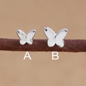 May include: Two silver butterfly stud earrings, labeled A and B, are displayed on a brown surface. The earrings are small, with a polished, reflective finish. The background is a solid, light brown color.