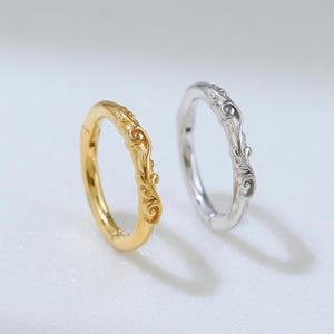May include: Two rings are shown, one gold and one silver. The gold ring has an elaborate, swirling pattern. The silver ring features a similar, yet more delicate, design. Both rings are set against a soft, neutral background, emphasising their intricate details.
