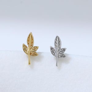 May include: A pair of leaf-shaped stud earrings. One earring is gold-colored, and the other is silver-colored. The earrings are small and detailed, with visible veins on the leaves. The background is a soft, white surface.