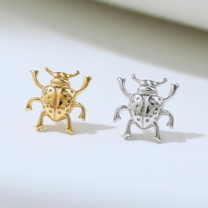 Puede incluir: Dos pendientes pequeños con forma de insecto. Uno es dorado, el otro plateado. Ambos tienen cuerpos detallados, antenas y patas. Los pendientes están sobre un fondo blanco.
