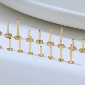 May include: A collection of gold-colored lip piercing studs of varying lengths and sizes. The studs have a flat, circular base and a straight post. The jewelry is arranged on a white surface, with a curved white object in the background.
