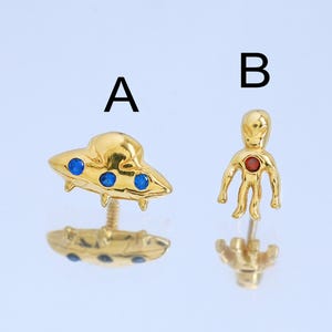 May include: Two gold-colored stud earrings. One earring is a UFO with blue accents, and the other is an alien figure with a red center. The earrings are on a reflective surface.