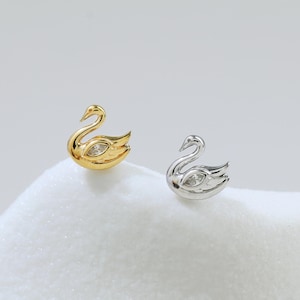 Könnte beinhalten: Zwei schwanförmige Ohrstecker. Einer ist goldfarben mit einem klaren Edelstein im Auge, der andere ist silberfarben mit einem klaren Edelstein im Auge. Die Ohrringe werden auf einer weißen, strukturierten Oberfläche präsentiert.