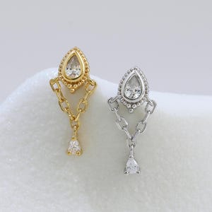 May include: Two stud earrings, one gold-toned and one silver-toned, each featuring a pear-shaped clear stone set in a beaded frame. Two delicate chains hang from the base of the setting, connecting to a small, dangling pear-shaped clear stone.