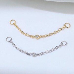 May include: Two delicate jewelry connector chains, one in a gold tone and one in a silver tone. Each chain features a small, circular, bezel-set clear stone at the center and a small jump ring at each end for attachment to other jewelry pieces.