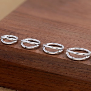 May include: A collection of silver-colored double-band rings of varying sizes, arranged on a dark wooden surface. The rings have a smooth, polished finish and a minimalist design. The rings are displayed in a row, showcasing their different sizes.