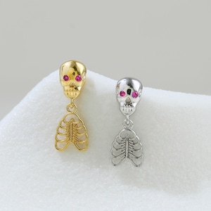 May include: A pair of earrings featuring a skull design with red accents for the eyes, and a rib cage pendant. One earring is gold-colored, and the other is silver-colored. The earrings are displayed on a white, textured surface.