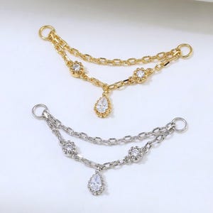 Può includere: Due connettori per catena decorativi, uno color oro e uno color argento. Ogni pezzo presenta due catene drappeggiate, due piccoli accenti circolari con pietre chiare incastonate e un ciondolo centrale a forma di goccia con una pietra chiara.