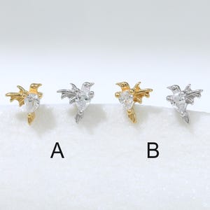 Pode incluir: Quatro pequenos brincos em forma de pássaros a voar com pedras transparentes em forma de lágrima no centro. Dois brincos são dourados e dois são prateados. As letras A e B estão posicionadas abaixo do segundo e quarto brinco, respetivamente.