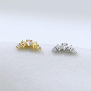 Könnte beinhalten: Zwei geschwungene Ohrstecker, einer in Gold- und einer in Silberoptik, jeweils mit einem zentralen, klaren Stein im Marquise-Schliff, flankiert von kleineren quadratischen und runden klaren Steinen in einem symmetrischen Kronendesign.