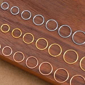 Könnte beinhalten: Eine Sammlung kleiner, kreisförmiger Ohrringe in verschiedenen Metallausführungen, darunter Silber, Gold und Roségold. Die Ohrringe sind auf einer dunklen Holzoberfläche angeordnet, die ihre unterschiedlichen Größen und Farben zeigt. Das Bild ist gut beleuchtet.