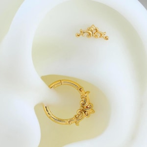 Può includere: Un set di gioielli per orecchie color oro composto da un piccolo perno decorativo curvo con intricati dettagli in filigrana e un anello a cerchio incernierato abbinato con un ornato design a volute su un lato del cerchio.