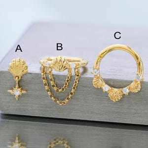 Puede incluir: Juego de joyas doradas con tres diseños distintos. Un pendiente con un dije de concha y estrella, un anillo con una concha y detalle de cadena, y un pendiente de aro con detalles de concha y pequeñas gemas. Todas las piezas se muestran sobre una superficie reflectante.