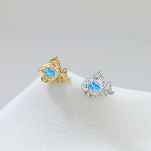 May include: Two fish-shaped earrings, one gold-toned and one silver-toned, each with a blue gemstone center. The earrings are set on a white surface. The fish design features detailed fins and scales.