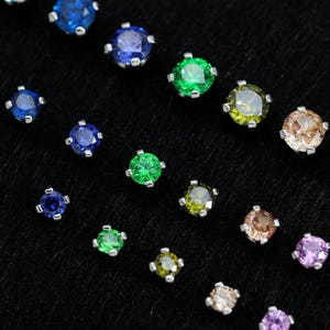 Pode incluir: Uma variedade de brincos de pressão com pedras preciosas em várias cores, incluindo azul, verde, amarelo, rosa e roxo. Cada brinco apresenta uma gema de corte redondo em uma moldura de metal prateado. Em um fundo preto.