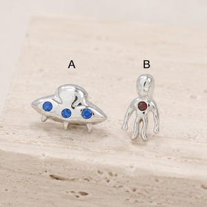 以下が含まれることがあります： 銀色の金属製スタッドピアス2点。ピアスAは3つの小さな青い円形の石が付いた空飛ぶ円盤の形をしています。ピアスBは胴体の中央に小さな赤い円形の石が付いたエイリアンの形をしています。