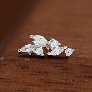 May include: A small, silver-toned earring with a cluster of clear, sparkling gemstones. The design features a mix of marquise and round-cut stones, arranged in a delicate, asymmetrical pattern. The earring is set against a warm, wooden surface.