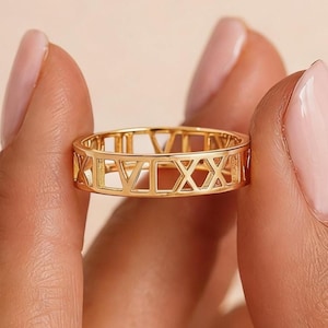 Pode incluir: Um anel dourado com um design de algarismos romanos. O anel é segurado entre dois dedos contra um fundo macio e neutro. O design do anel apresenta algarismos romanos recortados, criando uma estética aberta e arejada. O anel é uma joia.