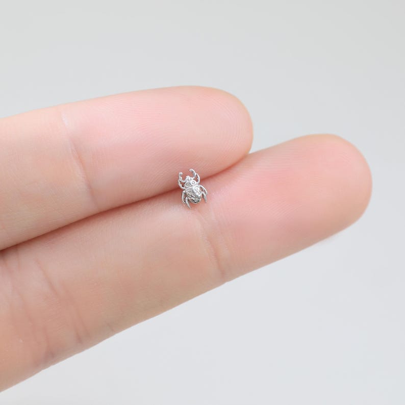 May include: A tiny silver-toned beetle stud earring with a metallic finish and intricate textured detailing on the shell and legs. The small insect-shaped piece features a rounded body and six distinct legs, showcasing a polished surface.