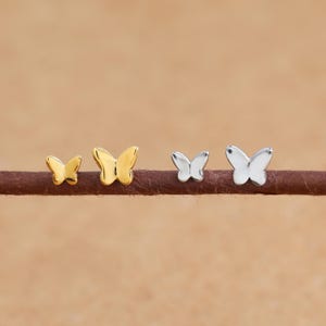 May include: Four butterfly stud earrings in gold and silver tones are displayed on a brown surface. The earrings vary in size, with two gold and two silver butterfly designs. The background is a solid, light brown colour.