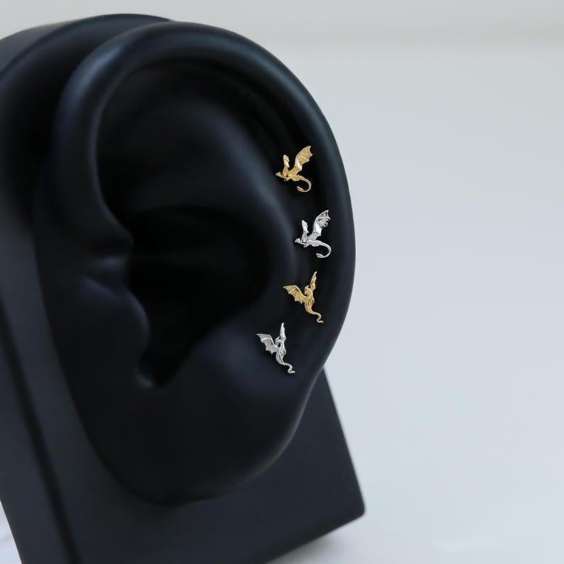 Op de afbeelding: Vier kleine oorknopjes in de vorm van een draak, verticaal gerangschikt langs het kraakbeen van een zwarte oordisplay. De set bevat twee goudkleurige en twee zilverkleurige drakenknopjes, elk met gedetailleerde vleugels, een lichaam en een staart.