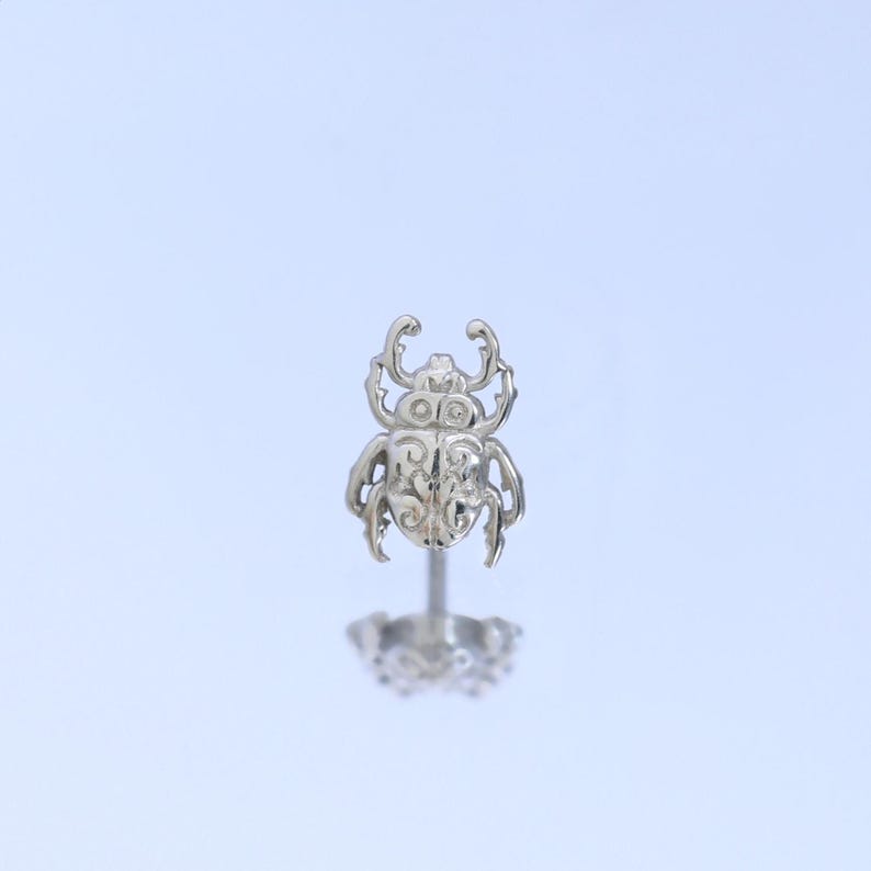 May include: A silver-toned stud earring featuring a detailed beetle design with intricate embossed scrollwork patterns on its back. The beetle has prominent mandibles and six legs, attached to a straight post with a matching silver-toned backing.