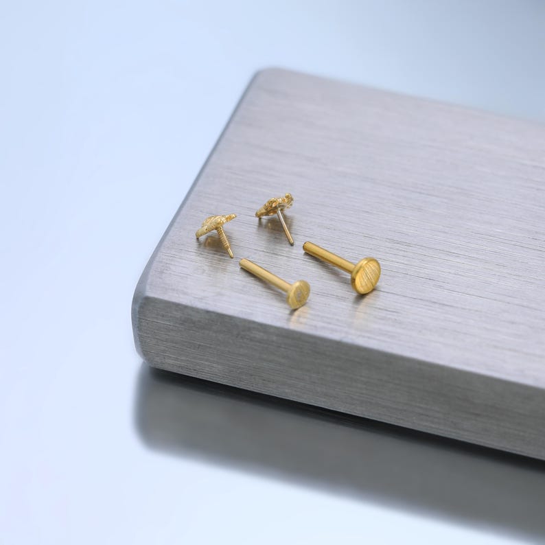May include: Two gold-toned stud earrings featuring a textured, star-shaped top and a flat, circular back post. The earrings are displayed with the post stems visible, showing the metallic finish and the distinct shape of the decorative front elements.