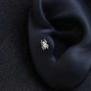 May include: A small, silver-colored beetle-shaped stud earring featuring intricate, embossed detailing on its surface. The metallic insect design is depicted with visible legs, antennae, and a rounded body, crafted as a singular piece of jewelry.