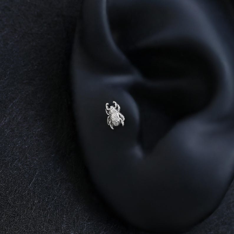 May include: A small, silver-colored beetle-shaped stud earring featuring intricate, embossed detailing on its surface. The metallic insect design is depicted with visible legs, antennae, and a rounded body, crafted as a singular piece of jewelry.