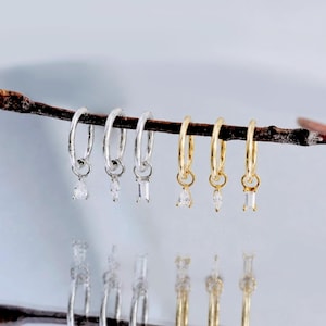 Puede incluir: Seis pendientes de aro pequeños colgados de una rama marrón fina. Tres aros plateados tienen piedras transparentes colgantes en forma de lágrima, marquesa y baguette. Tres aros dorados también tienen piedras transparentes colgantes iguales.