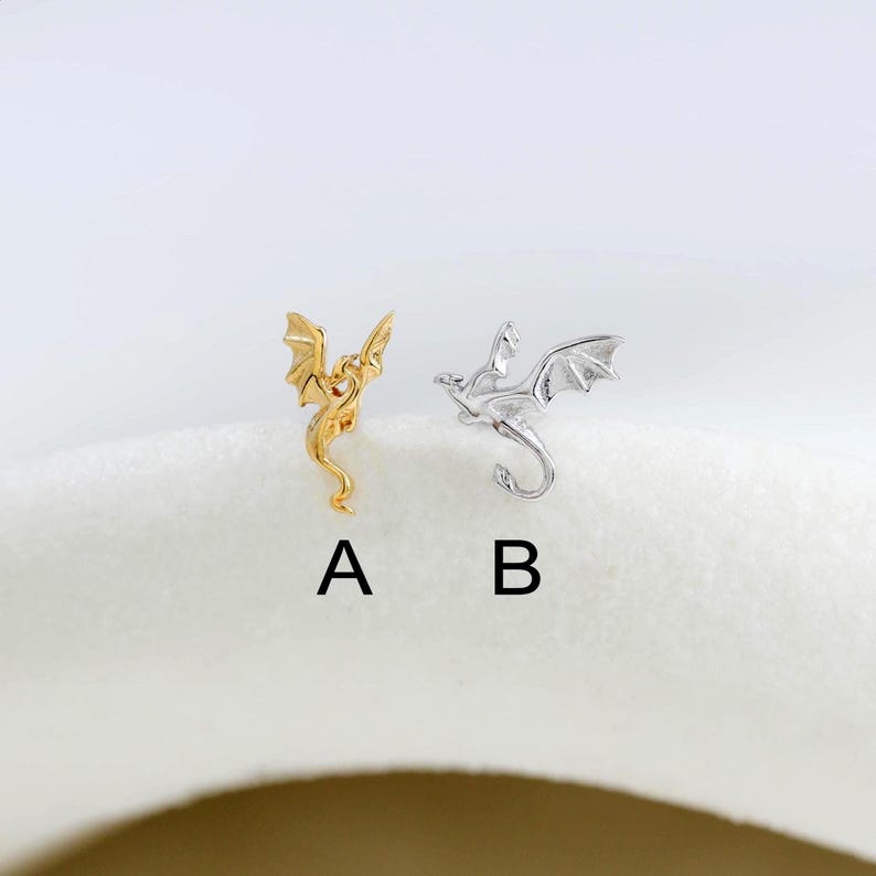 May include: Two dragon-shaped earrings. One is gold-colored, and the other is silver-colored. The dragons have outstretched wings and curved tails. The earrings are labeled A and B.