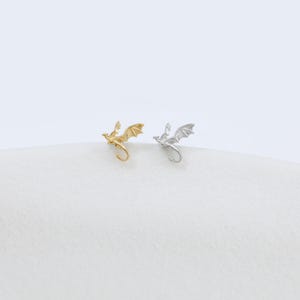 Op de afbeelding: Twee kleine oorknopjes in de vorm van vliegende draken. De ene oorbel heeft een metallic gouden afwerking, terwijl de andere een metallic zilveren afwerking heeft. Elke draak is afgebeeld in een vlieghouding met gespreide vleugels en een gekrulde staart.