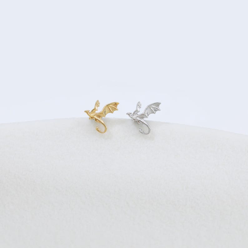 Op de afbeelding: Twee kleine oorknopjes in de vorm van vliegende draken. De ene oorbel heeft een metallic gouden afwerking, terwijl de andere een metallic zilveren afwerking heeft. Elke draak is afgebeeld in een vlieghouding met gespreide vleugels en een gekrulde staart.