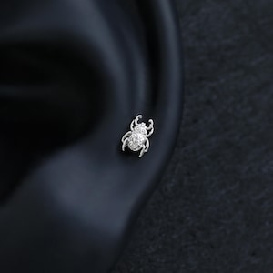 May include: A silver-toned stud earring shaped like a beetle. The insect design features intricate, embossed filigree patterns across its back, head, and legs. The metallic surface has a polished finish, highlighting the detailed texture of the beetle.