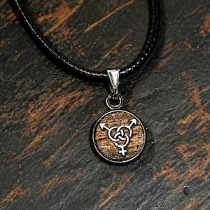 May include: A round pendant necklace with a wooden center and a silver-colored metal border. The pendant features a stylized symbol of interlocking arrows and a female symbol. The necklace has a black cord.